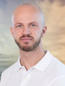 Professional head-and-shoulders portrait of a bald man with a beard in a white polo shirt, against a soft, sunset sky background.